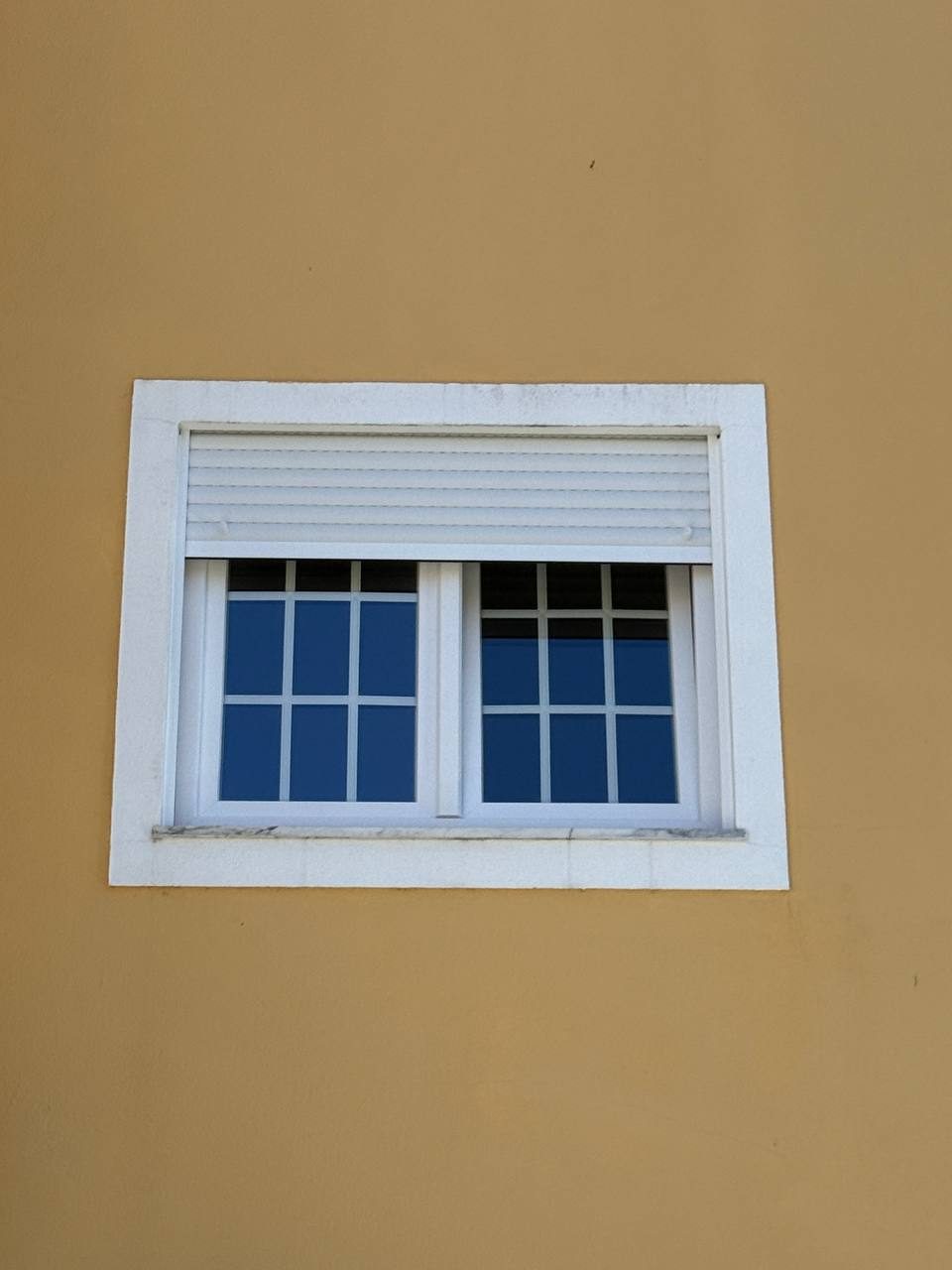 Janela em PVC branco com quadrícula e estore exterior, numa fachada amarela vista de fora