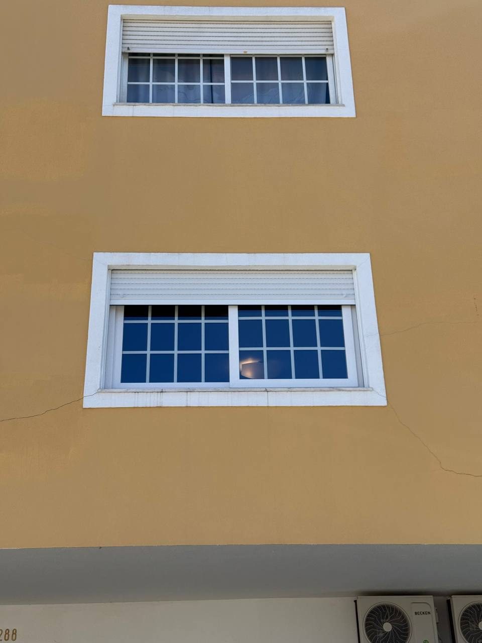 Duas janelas em PVC branco com quadrícula e estores exteriores, numa fachada amarela