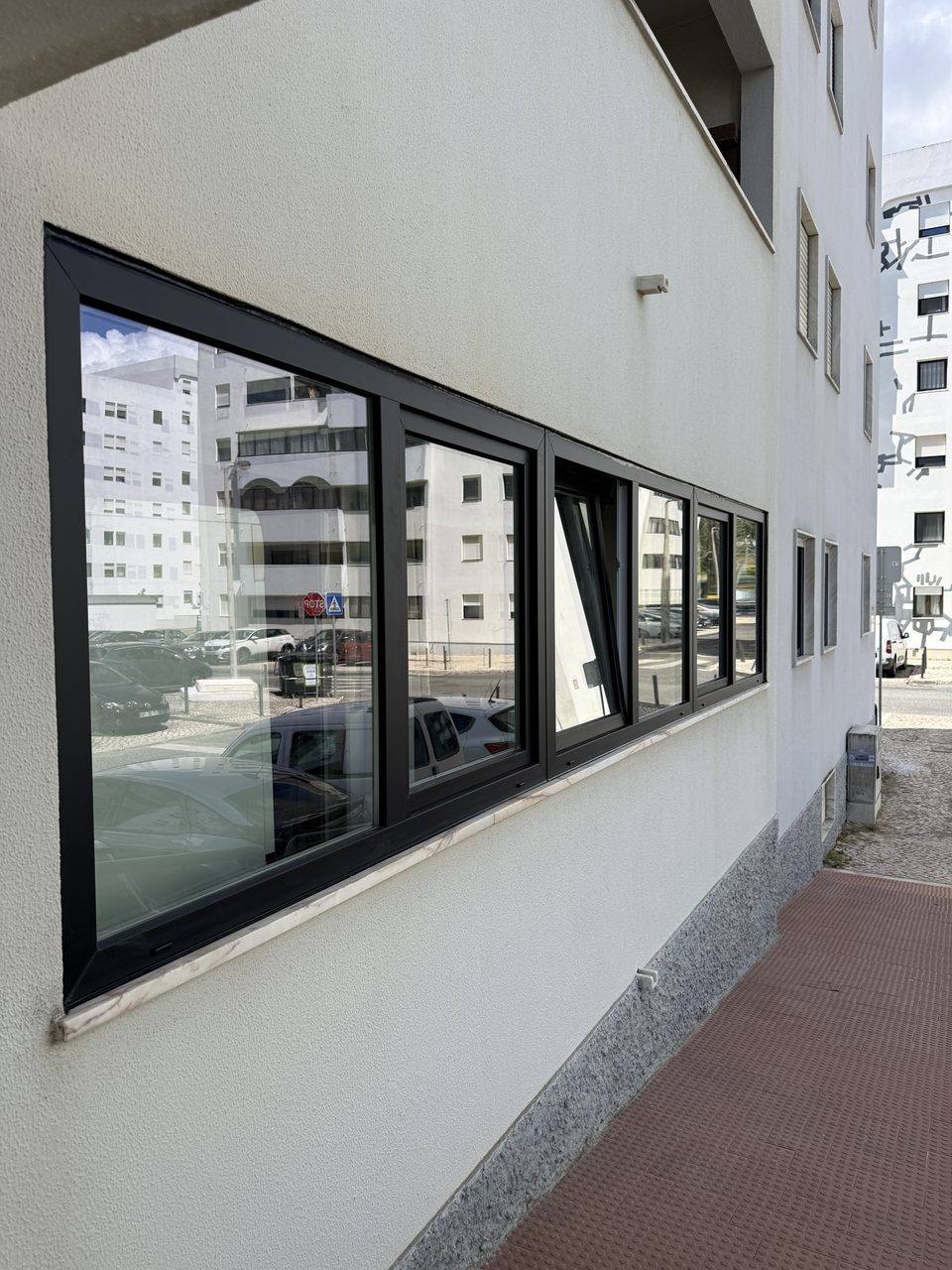 Janelas em PVC preto numa fachada branca, com uma folha basculante aberta, vistas da rua