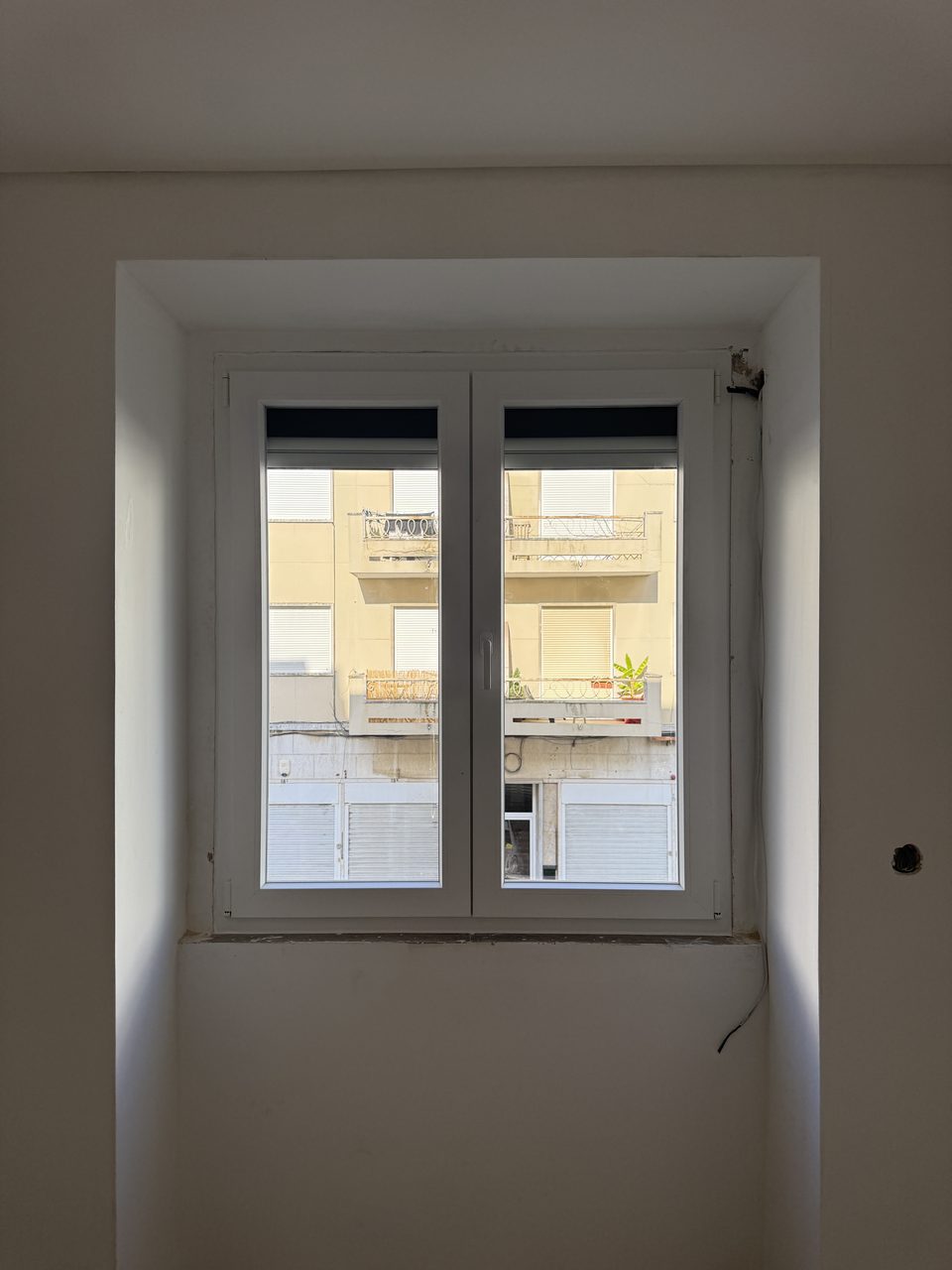 Janela de duas folhas em PVC branco num vão fundo, com o estore exterior no topo