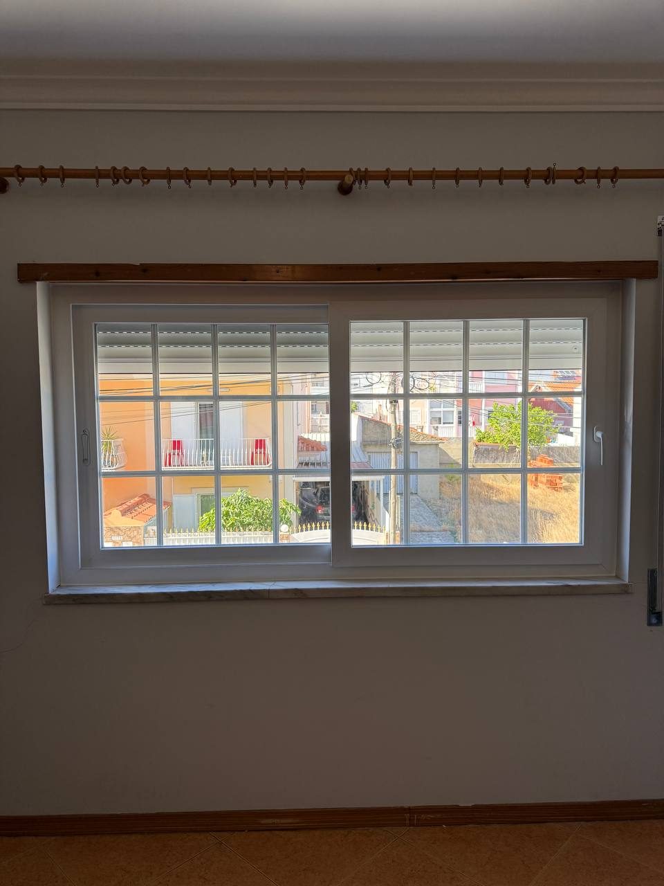 Janela larga de duas folhas em PVC branco com quadrícula numa sala, com o estore exterior visível no topo