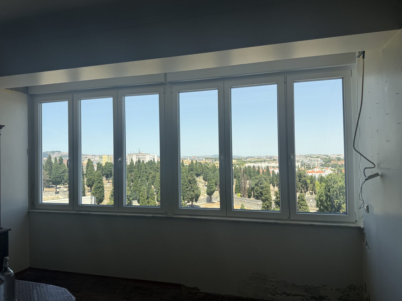 Janela de seis folhas em PVC branco ao longo de uma sala, com vista sobre árvores e a cidade