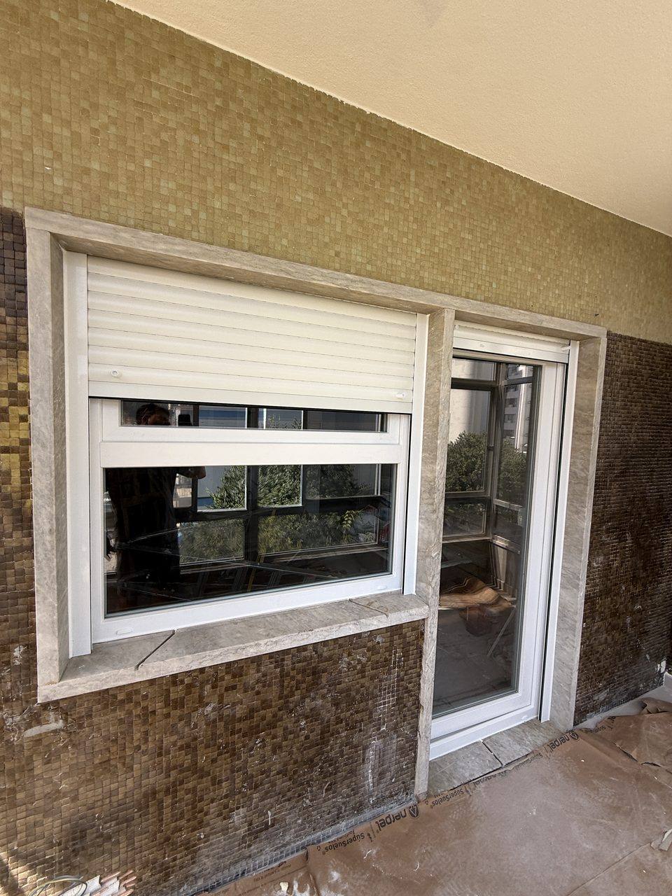 Janela em PVC branco com estore exterior e porta ao lado, numa varanda com pastilha cerâmica, durante a obra